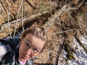 Beth looks skeptical in a selfie taken while standing on a fallen tree with a little bit of snow still on the ground.